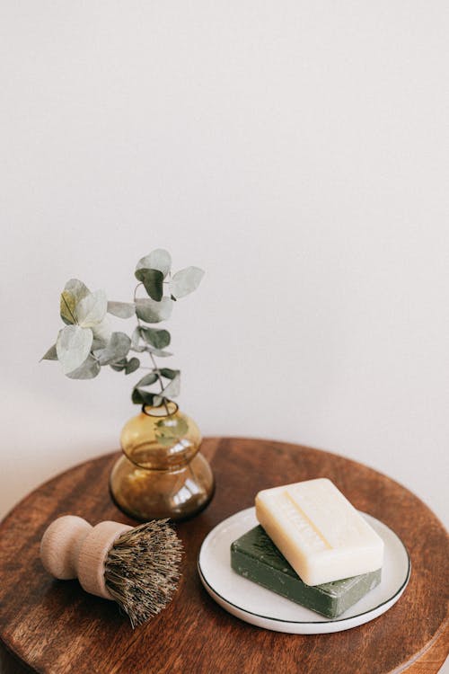 Free From above composition with assorted organic soaps on white ceramic holder and shaving brush with wooden handle near yellow transparent vase with plant on wooden round table against white wall Stock Photo